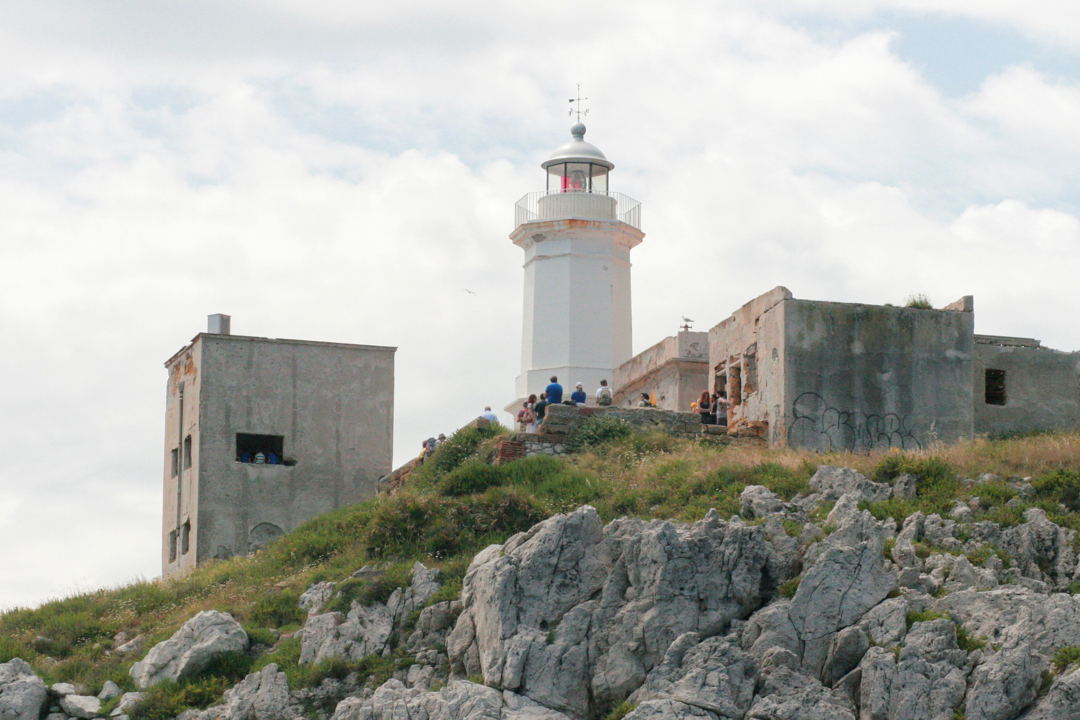 A lighthouse with a circular base and a dome on top, situated on a rocky hill with a grassy foreground.