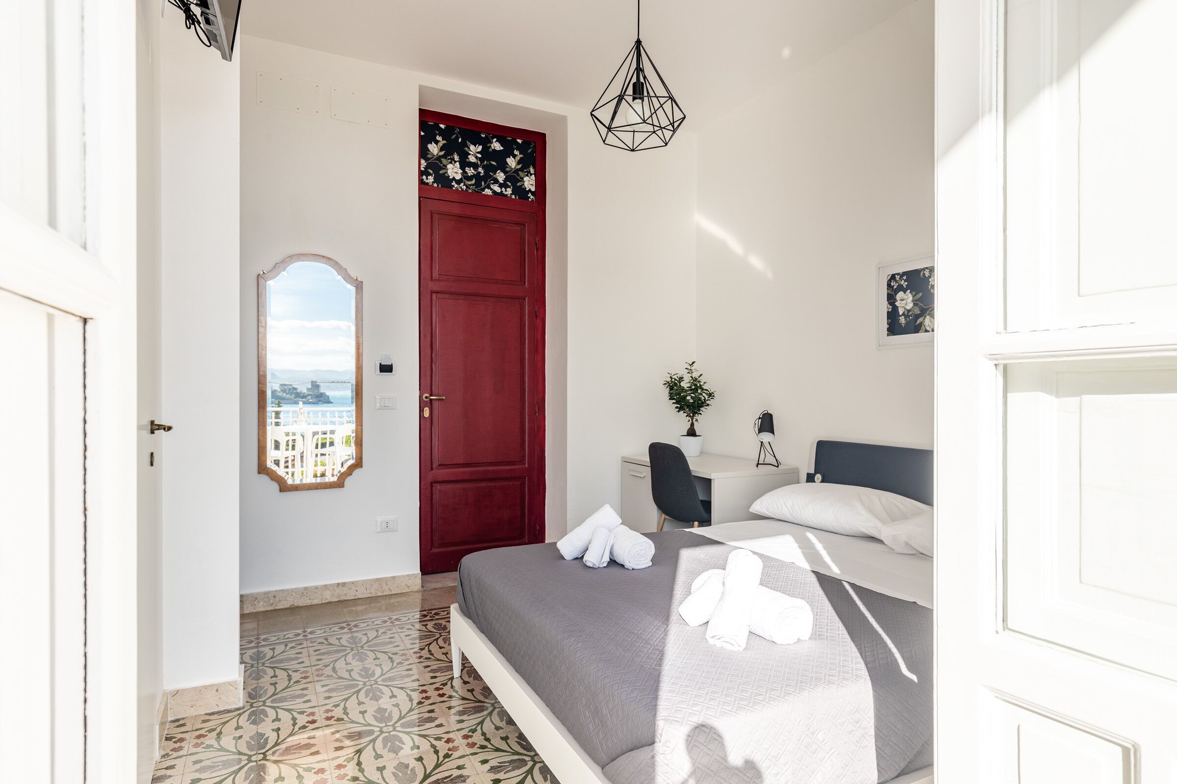 Un dormitorio con una cama, un escritorio, una silla, una puerta roja y vistas a través de una ventana.