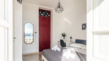 Un dormitorio con una cama, un escritorio, una silla, una puerta roja y vistas a través de una ventana.