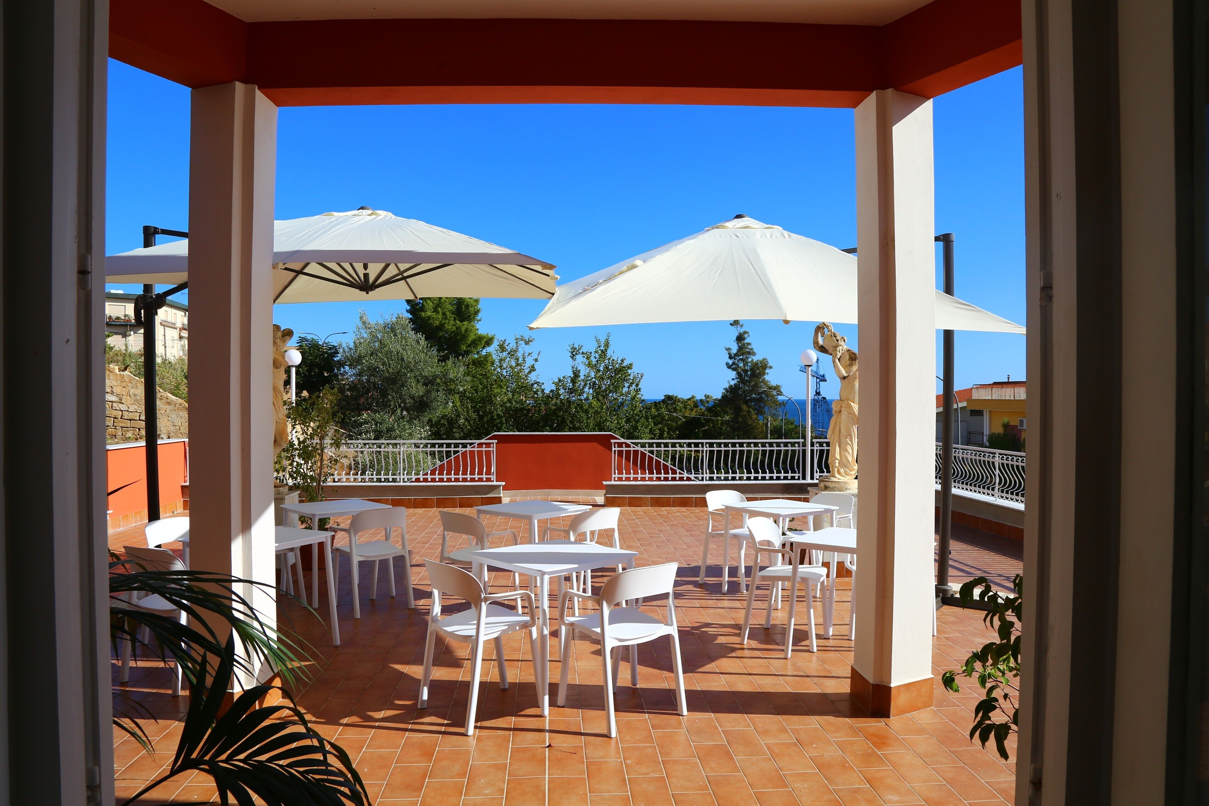 Une terrasse extérieure avec des tables et des chaises blanches, deux grands parasols blancs et une vue sur la mer.