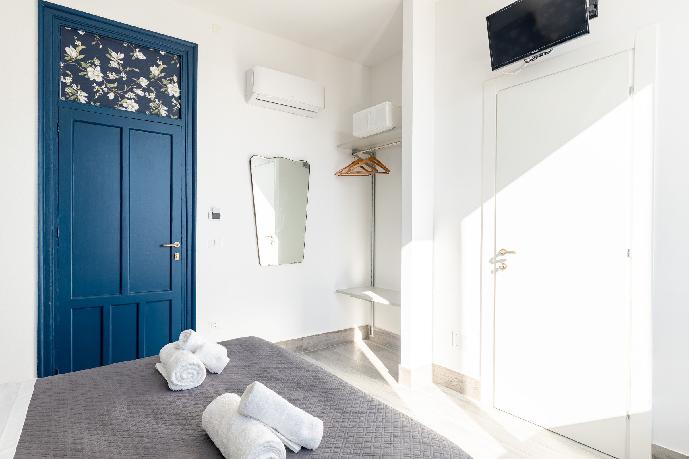 Une chambre d’hôtel avec une porte bleue, un lit recouvert d’une couverture grise, un miroir, un téléviseur fixé au mur et une armoire blanche.