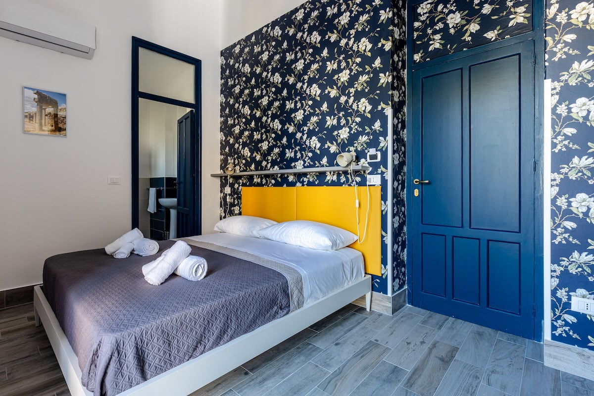 A bedroom with a bed, a mirror, a door, and a wall with floral wallpaper.