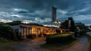 Un edificio con un espacio de estar al aire libre techado, un asta de bandera y un camino de entrada pavimentado.