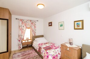 Ein Schlafzimmer mit Bett, Nachttisch, Kommode und einem Fenster mit Vorhängen. Drei gerahmte Bilder hängen an der Wand.