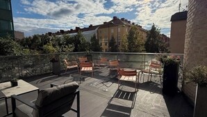Een terras op het dak met buitenmeubilair, uitzicht op gebouwen en een gedeeltelijk bewolkte lucht.