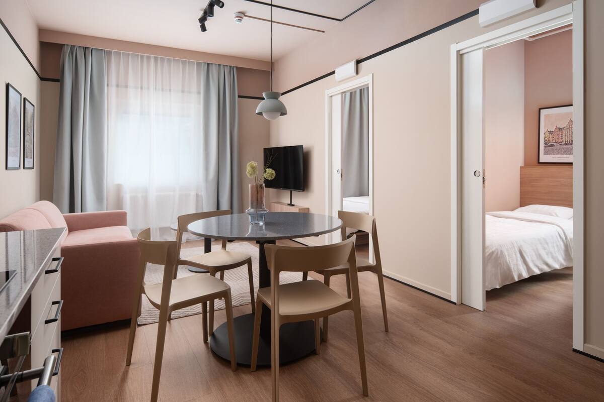 A modern hotel room with a round dining table, beige chairs, a sofa, a bed, and a TV on the wall.