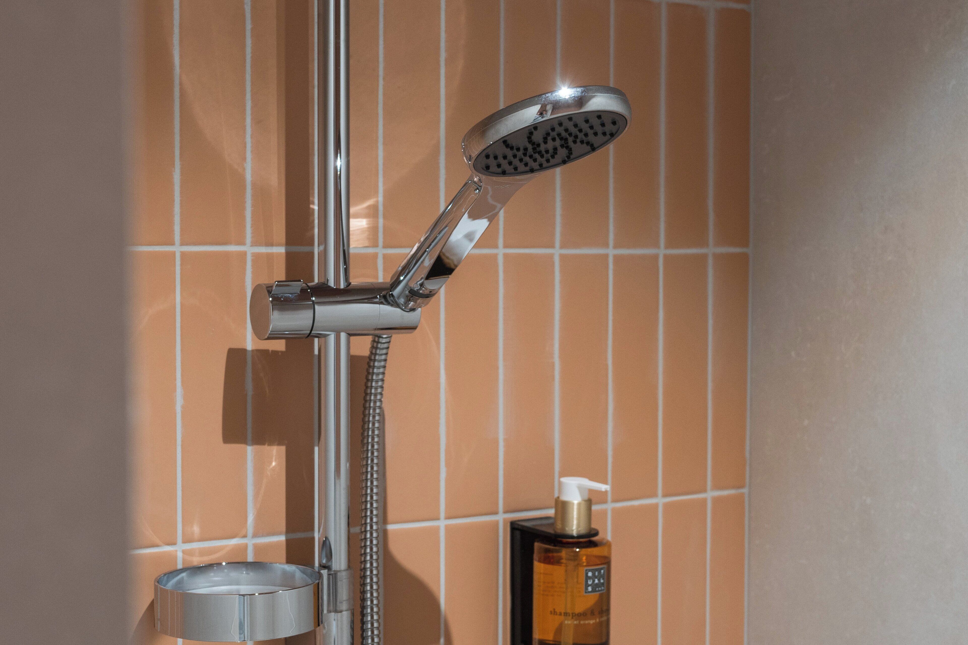 Un pommeau de douche muni d’un embout amovible, d’un tuyau flexible et d’une bouteille de shampoing, le tout fixé sur un mur carrelé.