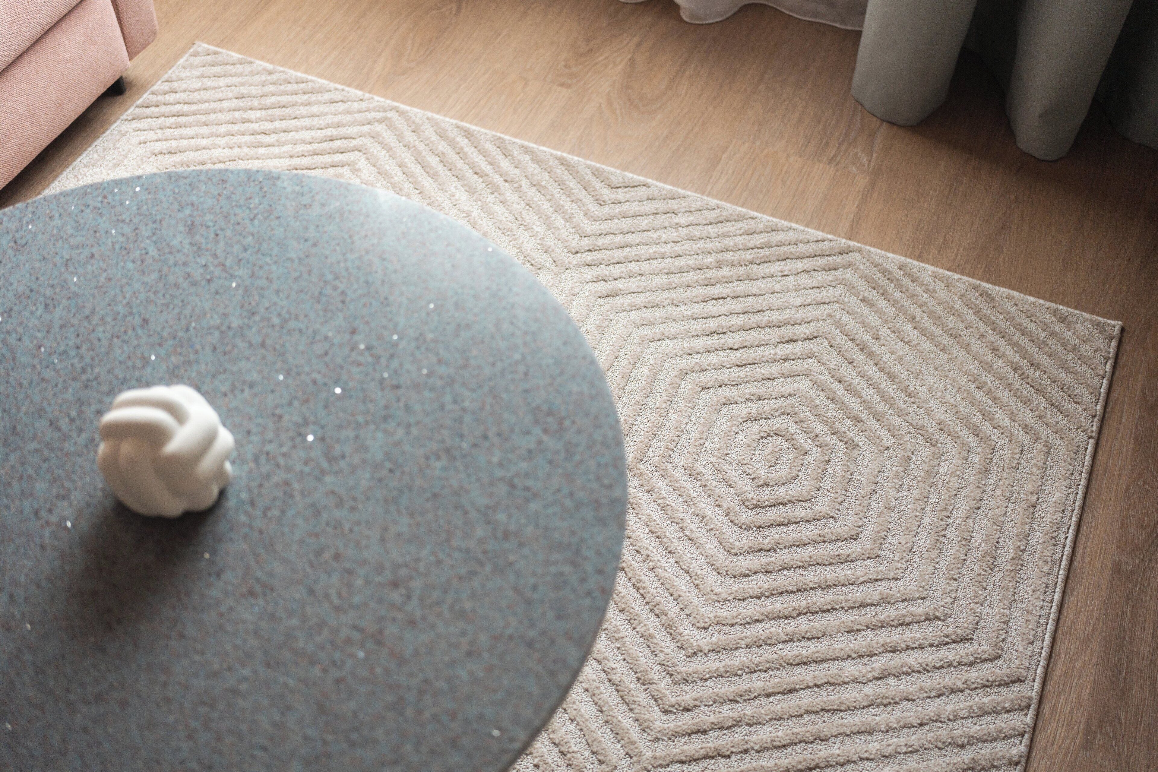 Una habitación con suelo de madera, una alfombra beige con diseño geométrico y una mesa redonda con un objeto blanco encima.