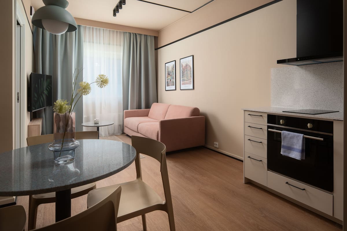 A modern living room with a round dining table, a sofa, a kitchen area with an oven, and a TV on the wall.