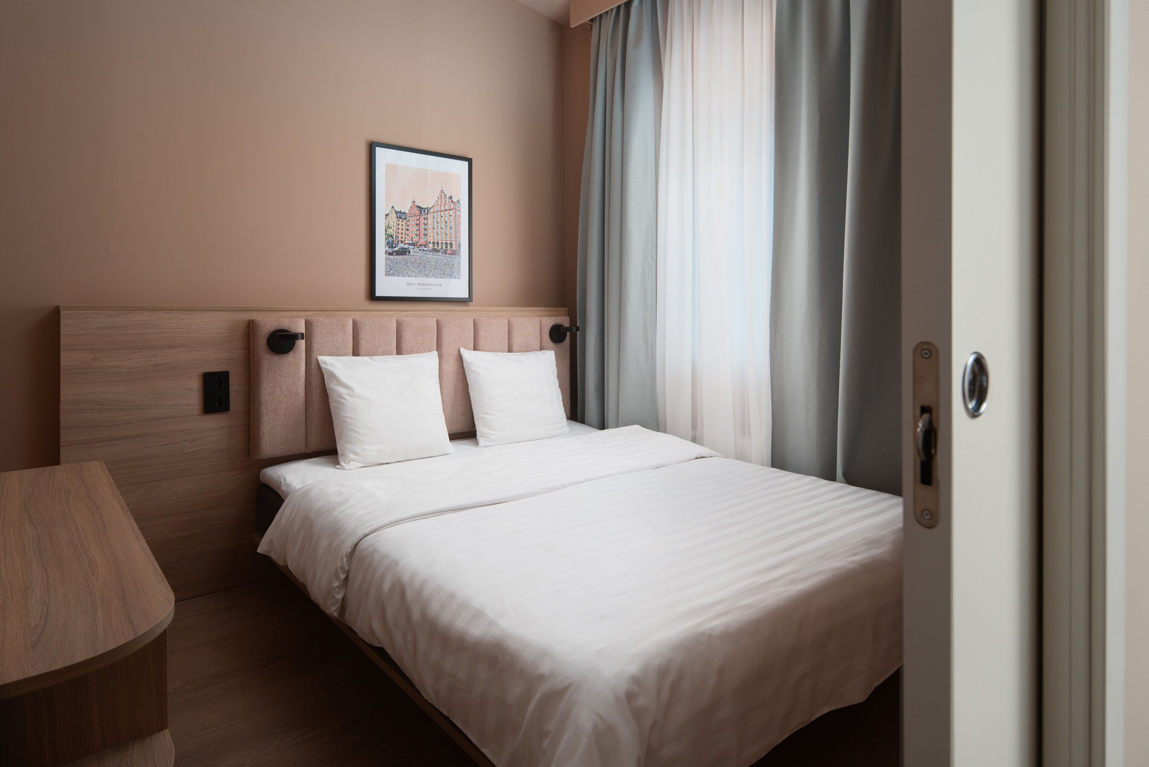 A hotel room with a bed, a bedside table, a framed picture on the wall, and a door with a handle.