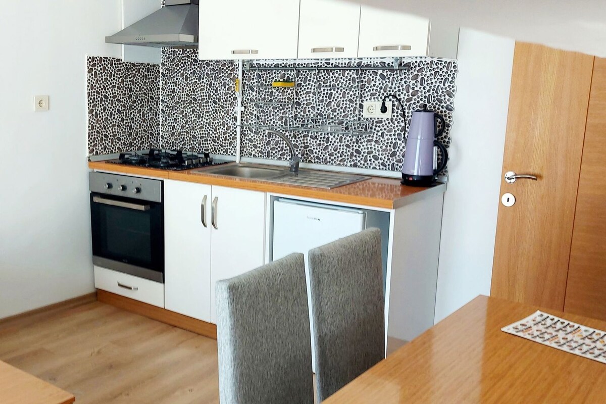 A compact kitchen with white cabinets, a built-in oven, and a microwave. There's a grey sofa with a blue cushion, a wooden dining table, and a grey chair.