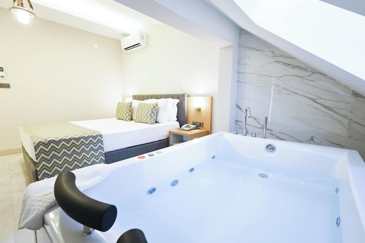 Ein modernes Hotelzimmer mit einer großen Badewanne, einem Bett mit gemusterten Betttextilien und einem Nachttisch mit Telefon und Lampe.