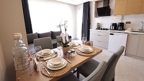 En matplats med ett träbord dukat för fyra personer, ett kök i bakgrunden och en soffa med en vas med vita blommor.