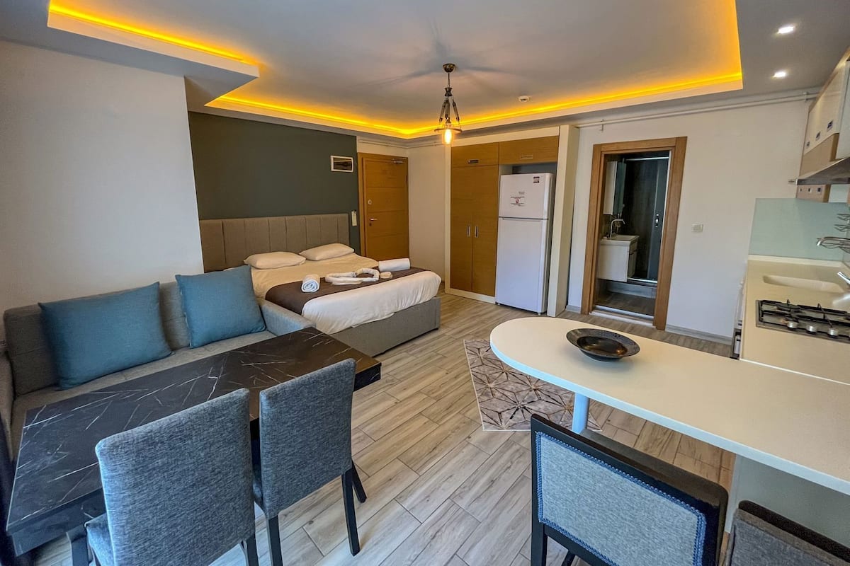 Ein modernes Hotelzimmer mit Bett, Sofa, Esstisch, Stühlen, Küchenzeile und einer Deckenleuchte.