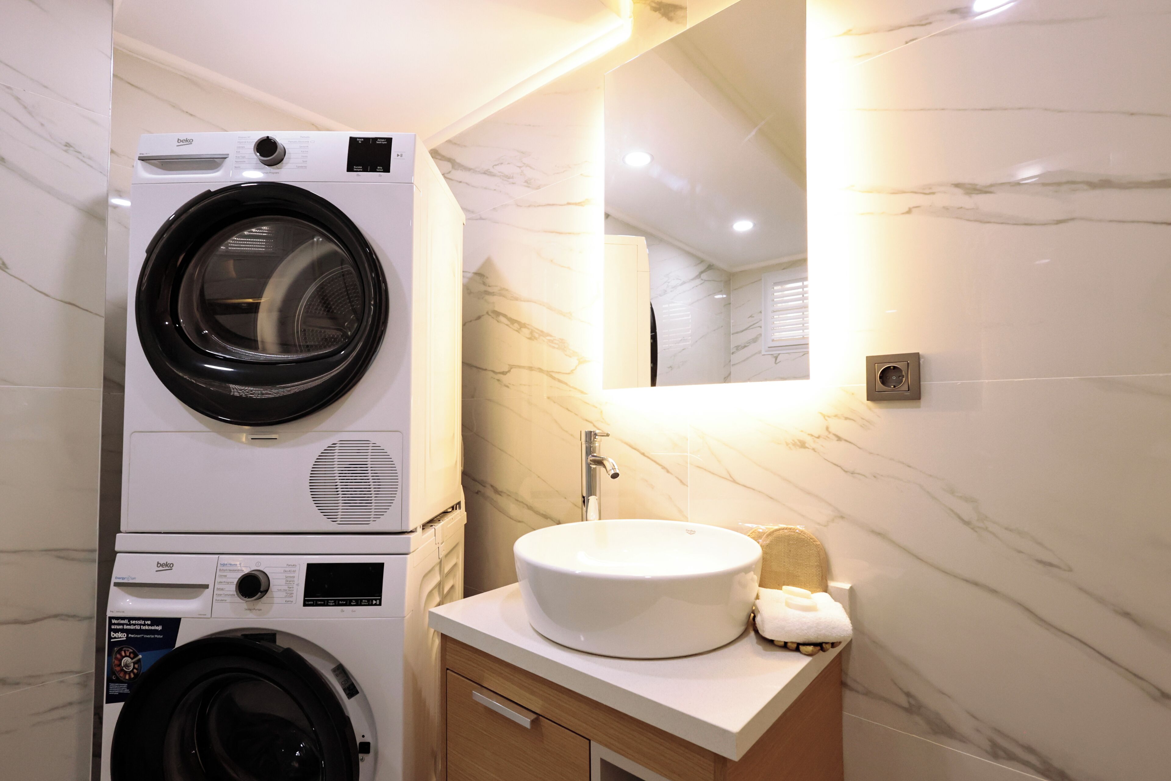 A compact laundry area with a front-loading washing machine and dryer, a sink with a marble countertop, and a mirror above the sink.