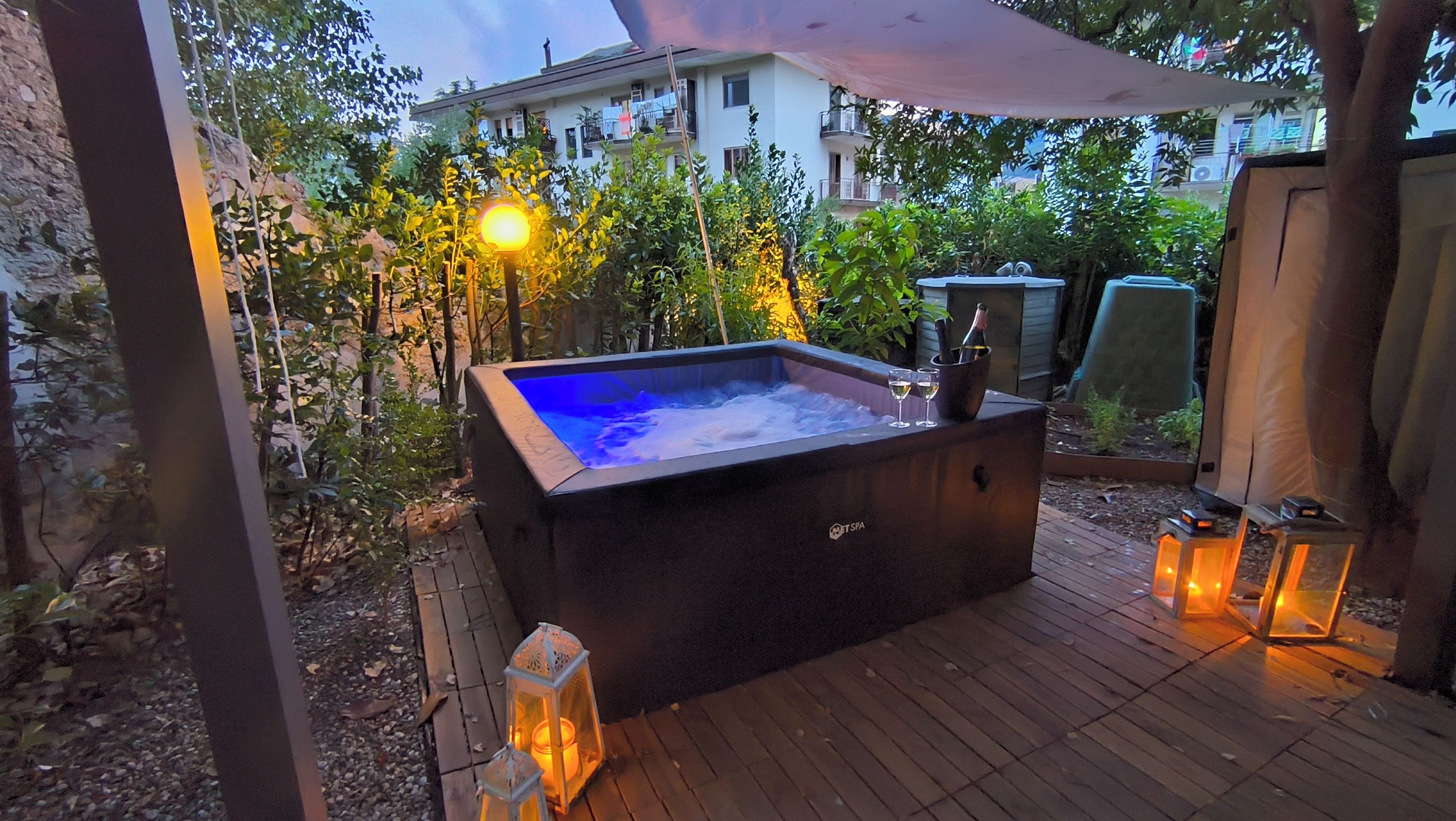 An outdoor hot tub with a wooden deck, surrounded by greenery and a building in the background.