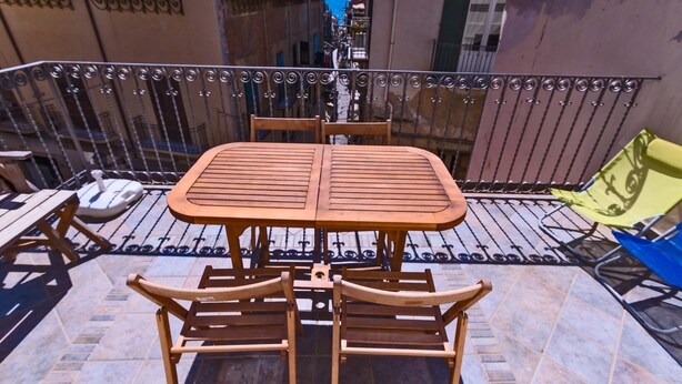 Una mesa y sillas de madera en un balcón con barandilla metálica, con vista a una calle estrecha.