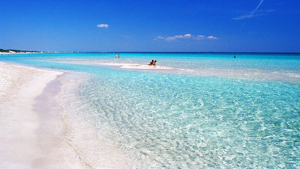 Une plage avec une eau turquoise et limpide, du sable blanc et quelques personnes au loin.