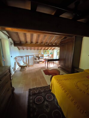 Une pièce avec un plafond en bois, un piano, une table à manger avec des chaises, un fauteuil en osier et un lit recouvert d’une couverture jaune.
