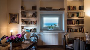 A cozy room with a view of the sea through a window, a bookshelf filled with books, a dining table with chairs, and a decorative vase with flowers. - Residence Torremuzza - Charming House In The Heart Of Palermo With Lovely View (Palermo)