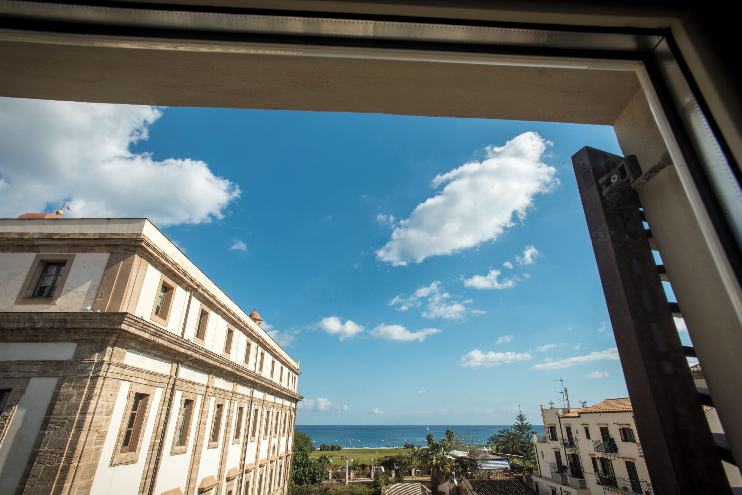 Aussicht aus einem Fenster auf eine Küstenlandschaft mit einem Gebäude im klassizistischen Stil und einem klaren Himmel mit Wolken.