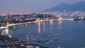 Un paysage urbain côtier, avec une montagne imposante en arrière-plan, une marina où sont amarrés de nombreux bateaux et une promenade en bord de mer bien éclairée.