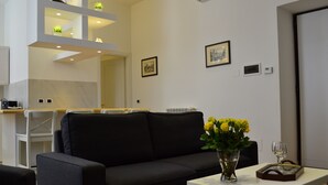 A modern living room with a black sofa, a small white table with a vase of yellow flowers, and a glass of wine. - Ursa Major - Gioligio Rome Central Luxury Suite (Rome)