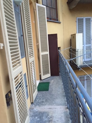 Um corredor externo estreito com grades de metal, um tapete verde na entrada e uma porta que dá para um edifício.