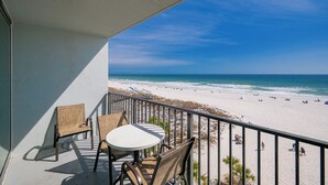 Balcón con sillas de mimbre y una mesa con vista a una playa donde hay gente y sombrillas.