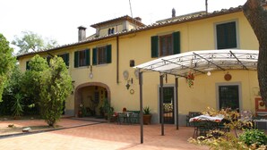 A yellow building with green shutters, a covered patio area with tables and chairs, and a brick pathway. - Villa Firenze Casa Serena (Montespertoli)