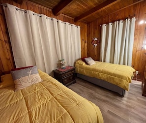 A wooden-paneled room with two beds, each with a yellow bedspread and a pillow with an anchor design. There is a nightstand with a lamp and a small decorative item.