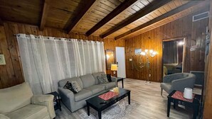 A cozy living room with a sofa, armchair, and coffee table, wooden walls, and a ceiling with exposed beams.