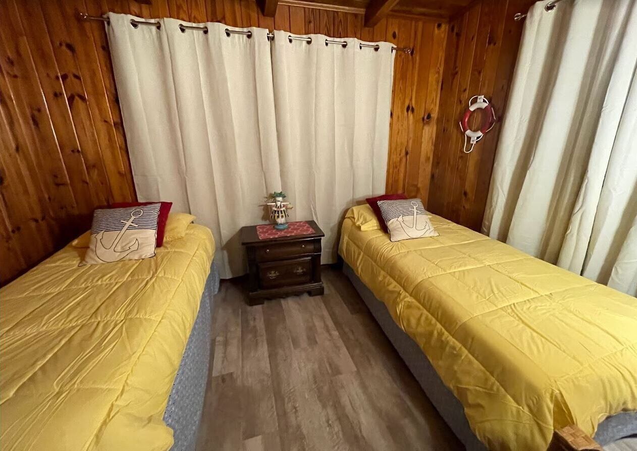 A room with two beds, wooden walls, a small table with a lamp, and a curtain.