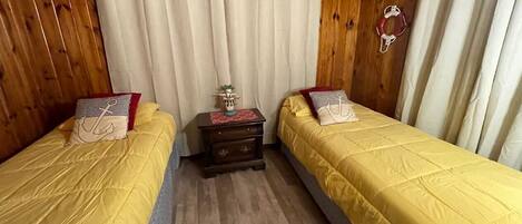 A room with two beds, wooden walls, a small table with a lamp, and a curtain.
