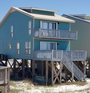 A two-story beachfront house with a balcony on the upper floor, wooden deck, and a staircase leading to the entrance.