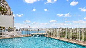 A swimming pool with a waterfall feature, surrounded by a white fence and a view of a building.