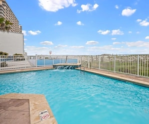 A swimming pool with a waterfall feature, surrounded by a white fence and a view of a building.