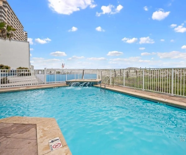 En swimmingpool med et vandfald, omgivet af et hvidt hegn og udsigt til en bygning.