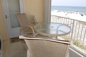 En balkon med et rundt glasbord og to fletstole, der ud til en strand med parasoller og mennesker.