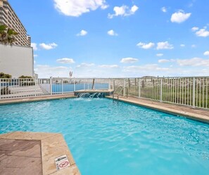 A swimming pool with a waterfall feature, surrounded by a white fence and a view of a building. - Low Density Gulf Front Condo | Sleeps 6 (Gulf Shores)
