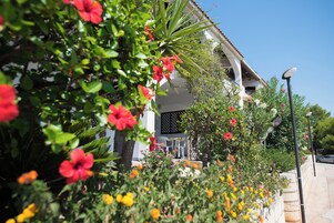 Un edificio con fachada blanca, un balcón con sillas y un jardín con diversas flores.