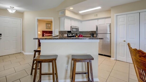 A kitchen with white cabinets, a central island with stools, stainless steel appliances, and a tiled floor.