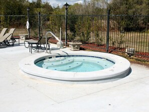 Un jacuzzi rond, dont le bord est recouvert de carreaux, entouré d’une terrasse en béton. On y trouve également des chaises longues et une petite table à proximité.