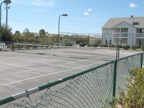 A tennis court with a chain-link fence, a bench, and a building in the background. - 2BR Gulf Shores Condo | Central Location + Short Drive to Beach (Gulf Shores)
