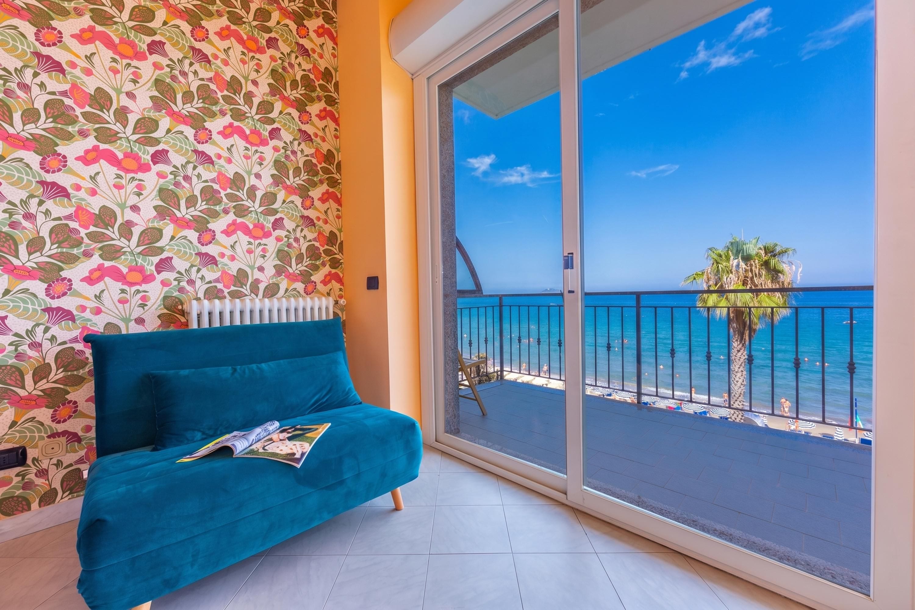 A room with a teal sofa, a patterned wall, and a balcony with a view of the sea.