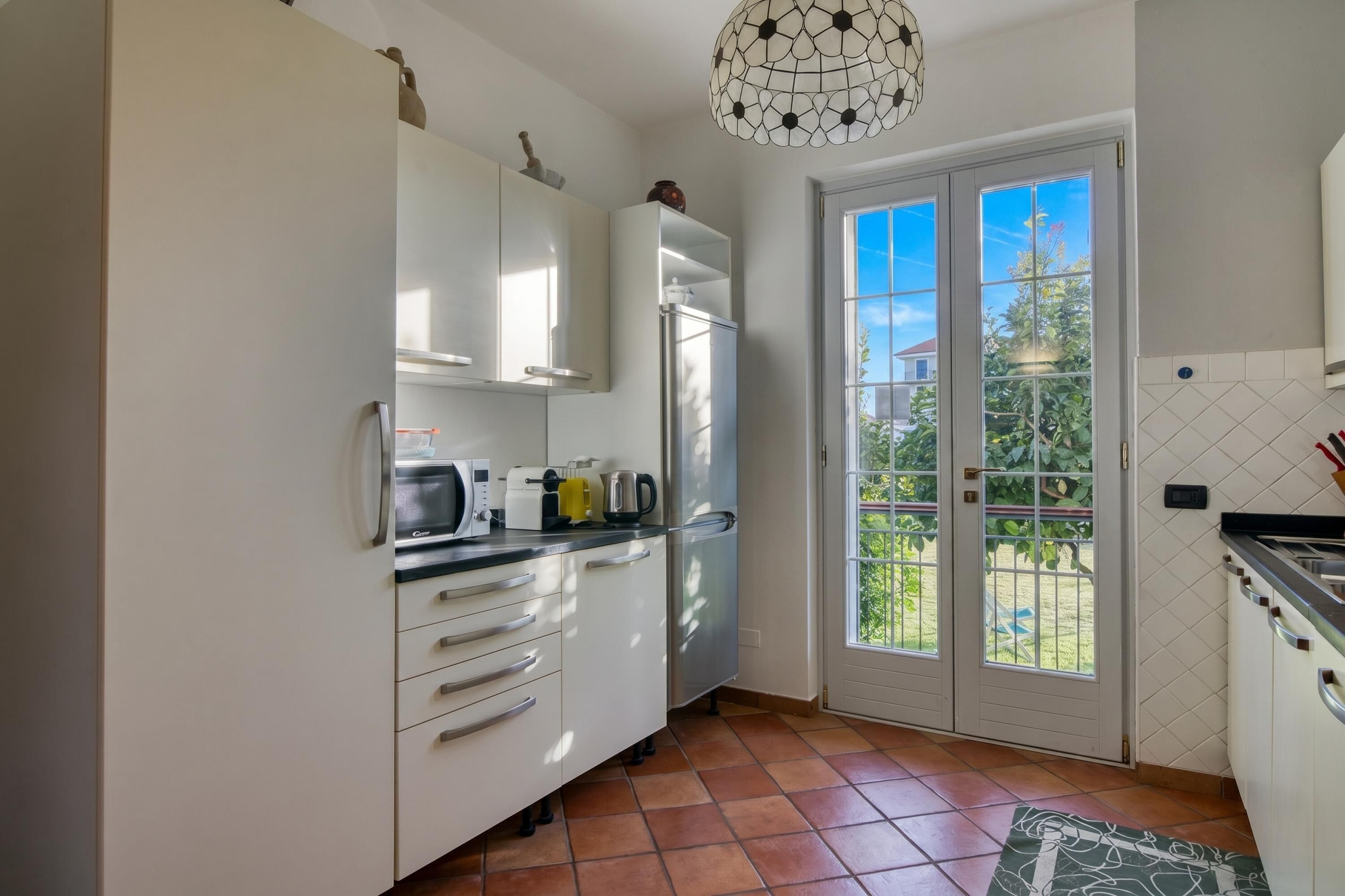 A kitchen with white cabinets, a refrigerator, a microwave, and a coffee maker. There is a large window with a view of greenery outside.