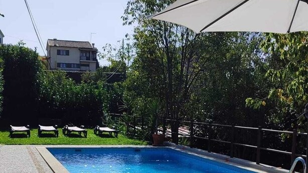 A swimming pool with a tiled deck, surrounded by greenery and a building in the background.
