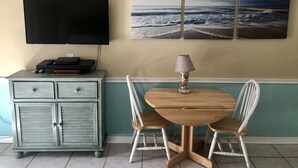 A dining area with a round wooden table, two white chairs, a lamp, and a television on a cabinet. - Sugar Beach 138 | Cozy Orange Beach Studio w Pool & Side Gulf View | Sleeps 4 (Orange Beach)