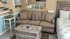 A cozy room with a beige sofa, a small white stool, a wooden coffee table, and a bed with a floral bedspread. - Sugar Beach 138 | Cozy Orange Beach Studio w Pool & Side Gulf View | Sleeps 4 (Orange Beach)