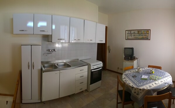 A compact kitchen with white cabinets, a double sink, and a small oven. There's a dining table with a patterned tablecloth and a few chairs. A television is mounted on a cabinet, and a painting hangs on the wall.